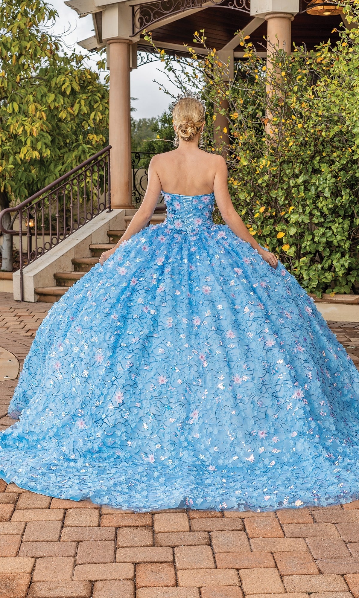 A woman with blonde hair in a low bun wears the Quinceanera Dress 1835 By Dancing Queen, a strapless ball gown with a floral pattern, standing on a brick patio facing away from the camera.
